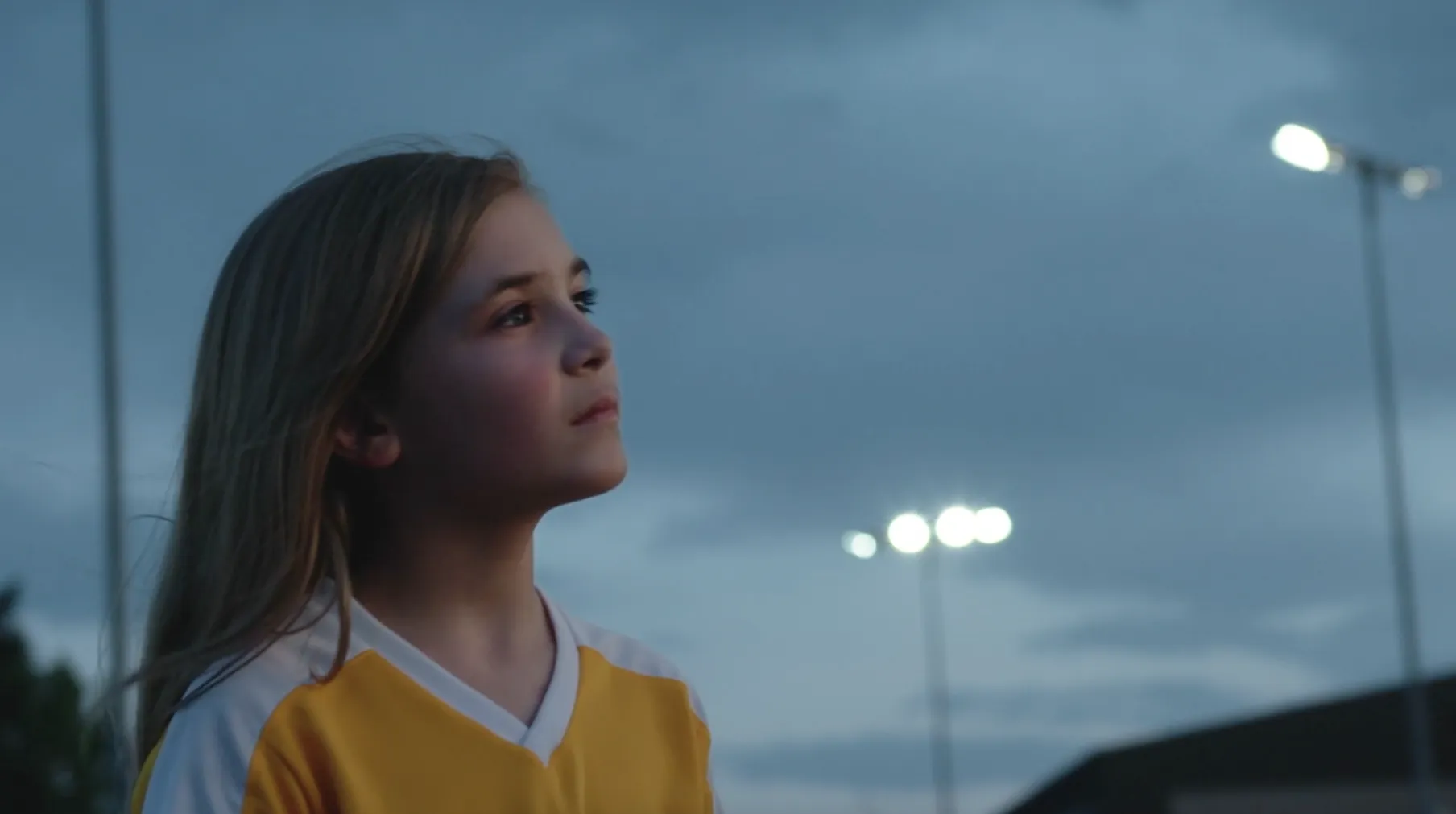 Girl in a yellow sports jersey looks up beneath stadium lights in My Time